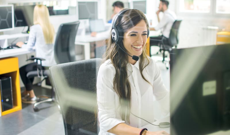Eine Frau, die ein Headset trägt, sitzt an einem Traderfox Trading-Desk und arbeitet an einem Computer in einem modernen Büro mit anderen Personen im Hintergrund.