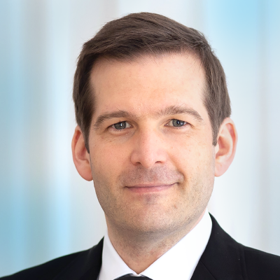 Mario Perl in schwarzem Anzug und weißem Hemd, der als einer der Referenten auftritt, blickt in die Kamera und lächelt leicht vor einem unscharfen blauen und weißen Hintergrund.