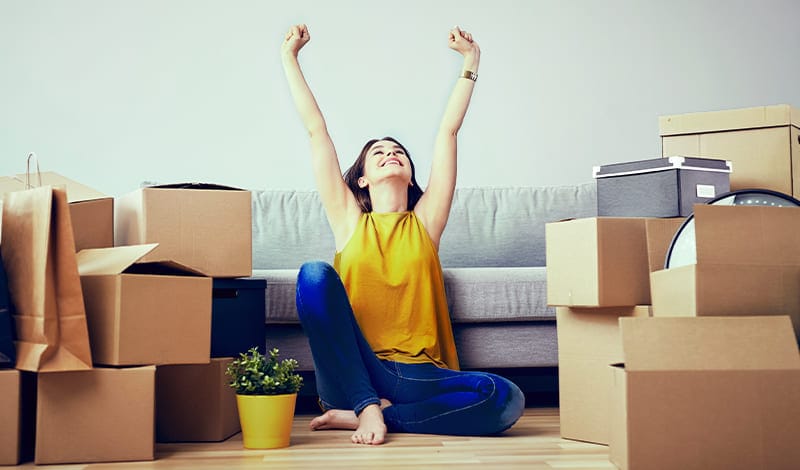 A woman sits on the floor with her arms raised, surrounded by moving boxes and a small potted plant, celebrating a successful deposit transfer in front of her sofa.