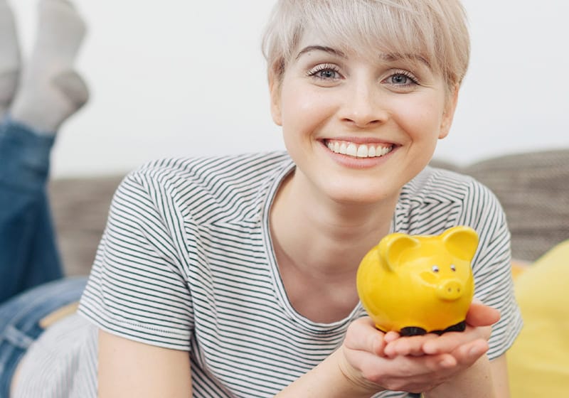 Smiling person with short blonde hair holds a yellow piggy bank while lying on a couch in a striped shirt, demonstrating the joy of saving with tradable products.