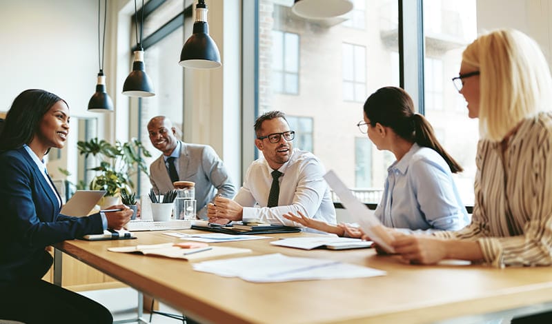 Five people in business attire sit around a conference table in a modern office and discuss their company portfolio; papers, laptops and documents lie on the table.