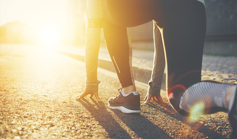 Eine Person in Sportkleidung hockt an der Startlinie auf einer sonnenbeschienenen Straße und bereitet sich auf den Lauf vor. Die Szene wird von hellem Sonnenlicht hinterleuchtet und fängt einen Moment der Konzentration und Energie ein - bereit zum Handeln, ähnlich wie nahtloser Zahlungsverkehr in Bewegung.
