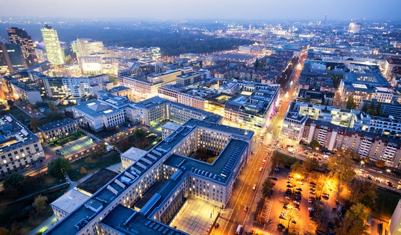 Aerial view of a city at dusk with illuminated streets, modern buildings ideal for real estate investment and scattered trees, with a large city park in the background.