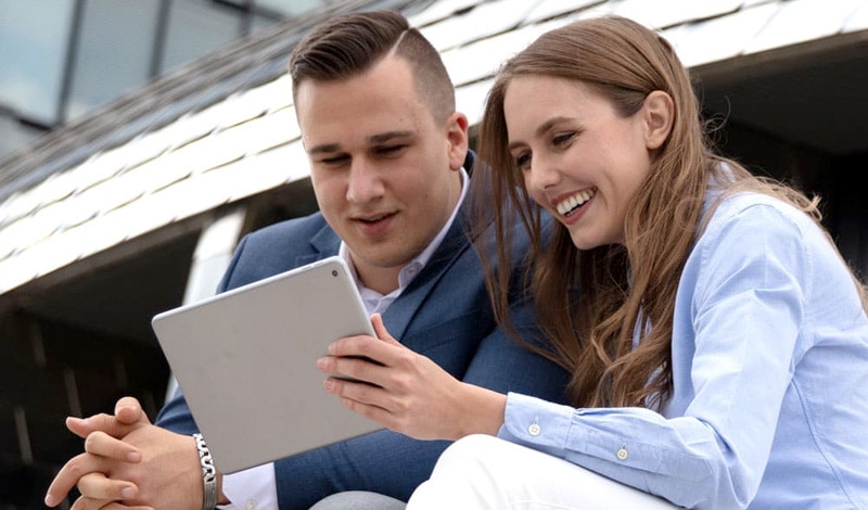 Ein Mann und eine Frau sitzen im Freien und lächeln, während sie auf ihrem Tablet Orderarten erkunden und mit CapTrader über Depot- und Handelsoptionen diskutieren.