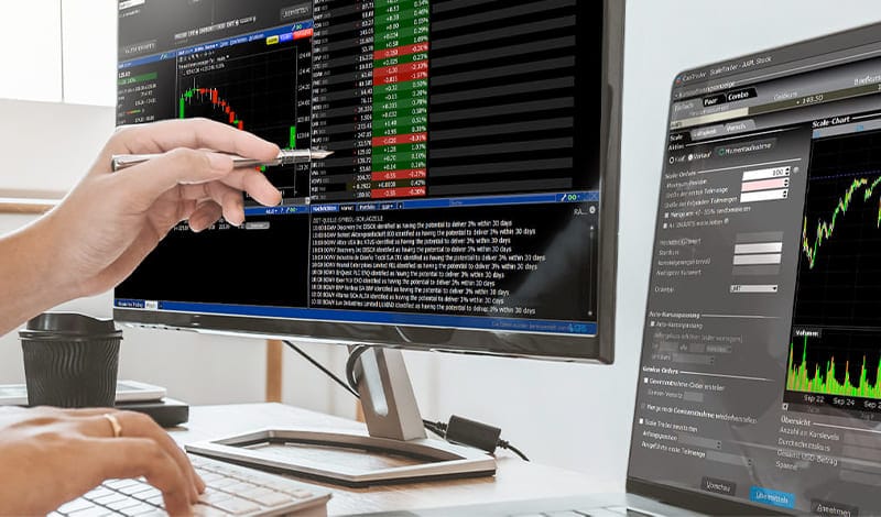 Person analyzing stock market data on two monitors at a trader workstation, with charts, news and financial information displayed; one hand points to the screen with a pen.