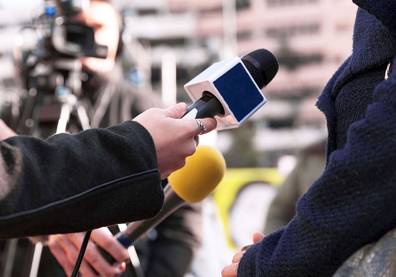Mehrere Personen halten Mikrofone in Richtung einer nicht identifizierten Person für Interviews im Freien, mit einer Kamera und unscharfen Gebäuden im Hintergrund - sie fangen spontane Momente ein, ähnlich wie die von CapTrader.