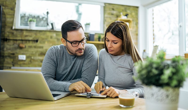 Ein Mann und eine Frau sitzen mit einem Laptop und Dokumenten an einem Tisch und diskutieren in einer gemütlichen Küchenumgebung über Papierkram und handelbare Produkte.