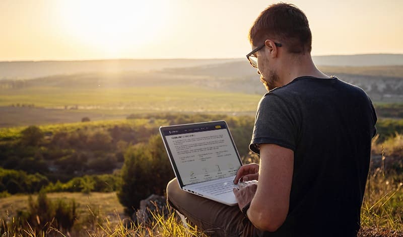 Eine Person sitzt bei Sonnenuntergang im Gras in der Nähe eines alten Depots und arbeitet an einem Laptop mit einer malerischen Landschaft im Hintergrund.