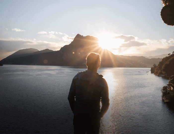 Eine Person steht an einem Gewässer und blickt auf die Berge, während die Sonne hinter ihr unter- oder aufgeht und den Himmel in ein helles Licht taucht - eine ideale Szene für eine inspirierende Imagebroschüre.