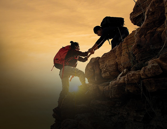 Zwei Wanderer mit Rucksäcken klettern bei Sonnenuntergang auf einen Felsvorsprung, wobei der eine dem anderen hinunter hilft - ein perfekter Moment von Teamwork und Abenteuer für Ihre nächste Imagebroschüre.
