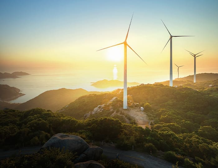 Drei Windturbinen stehen auf einem grünen Hügel bei Sonnenuntergang, mit Blick auf den Ozean und ferne Inseln unter einem klaren Himmel - ein ideales Motiv für eine Imagebroschüre, die Nachhaltigkeit und saubere Energie fördert.