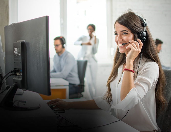 Eine Frau mit einem Headset sitzt lächelnd an einem Computer in einem Büro, während im Hintergrund andere Menschen arbeiten - eine ideale Szene für Ihre Imagebroschüre.