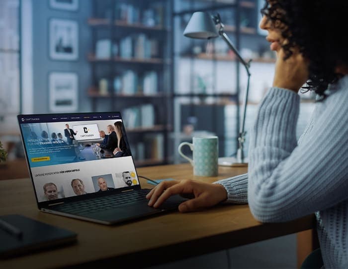 Eine Person sitzt an einem Schreibtisch in einem gemütlichen Raum und verwendet einen Laptop, auf dessen Bildschirm eine Imagebroschüre oder eine geschäftliche Website geöffnet ist. Auf dem Schreibtisch stehen eine Tasse und eine Lampe, im Hintergrund stehen Bücherregale.