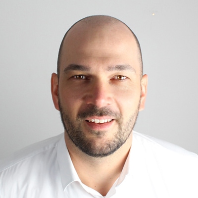 Referent Philipp Berger mit Glatze, kurzem Bart und Schnurrbart, der ein weißes Hemd mit Kragen trägt, blickt vor einem schlichten, hellen Hintergrund in die Kamera - das gelassene Erscheinungsbild eines professionellen Referenten.