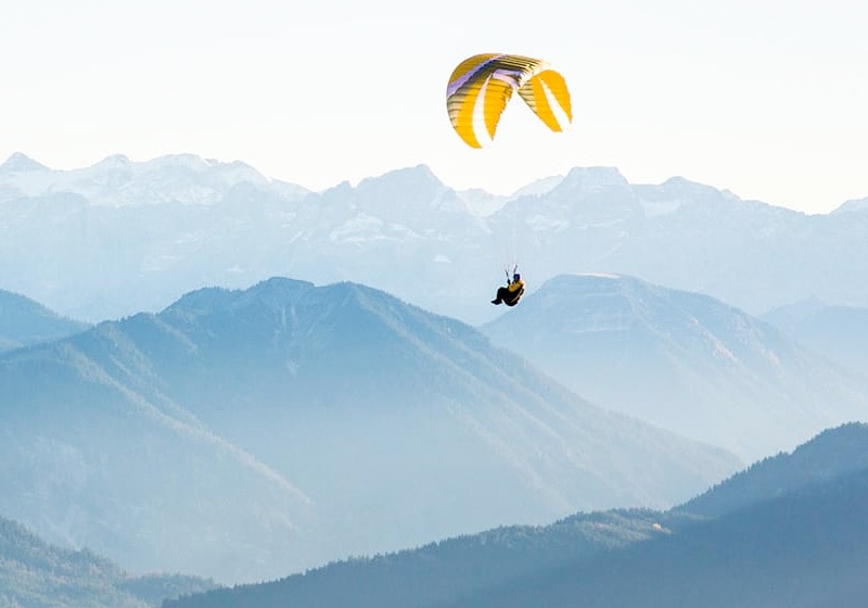 A person glides with a yellow and white parachute over mountainous terrain under clear skies, experiencing the same exhilaration and freedom as those who are actively involved.