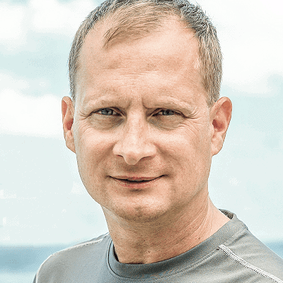 Ein Referent mittleren Alters mit kurzen hellbraunen Haaren und einem grauen Hemd blickt in die Kamera, während er im Freien vor einem verschwommenen Himmelshintergrund steht.