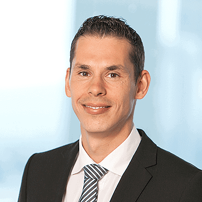 Andre Stagge in dunklem Anzug, weißem Hemd und gestreifter Krawatte posiert für ein professionelles Portraitfoto vor einem unscharfen hellblauen Hintergrund.
