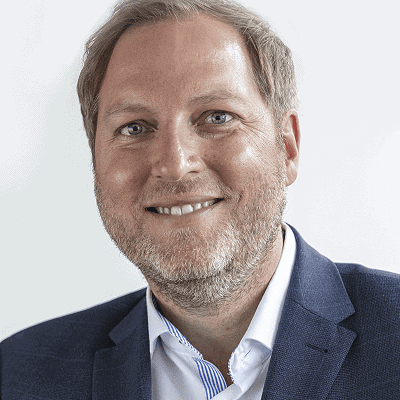 Tradingcoach Andreas Weiß mit hellbraunem Haar und Bart, der eine blaue Anzugsjacke und ein weißes Hemd trägt, lächelt vor einem schlichten Hintergrund in die Kamera - ein perfektes Bild für das professionelle Auftreten vieler Referenten.