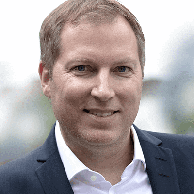 Geschäftsführer Christian Weiß mit kurzen hellbraunen Haaren, weißem Hemd und dunkler Anzugsjacke, lächelt in die Kamera, während er im Freien steht und das selbstbewusste Auftreten eines erfahrenen Referenten zeigt.