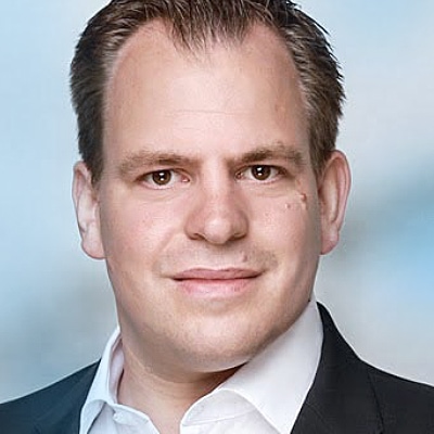 Tradingcoach Henning Henke in einer dunklen Anzugsjacke und einem weißen Hemd posiert für ein professionelles Foto als einer der Referenten vor einem unscharfen blauen Hintergrund.
