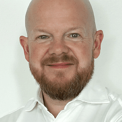 Tradingcoach Jens Rabe mit rötlich-braunem Bart und Schnurrbart lächelt in die Kamera. Er trägt ein weißes Hemd mit Kragen vor einem schlichten hellen Hintergrund.