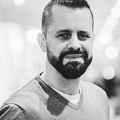 Luis Pazos mit kurzen Haaren und einem leichten Lächeln, möglicherweise einer der Referenten, trägt einen Pullover und steht in einem Innenraum. Das Bild ist in schwarz-weiß gehalten.