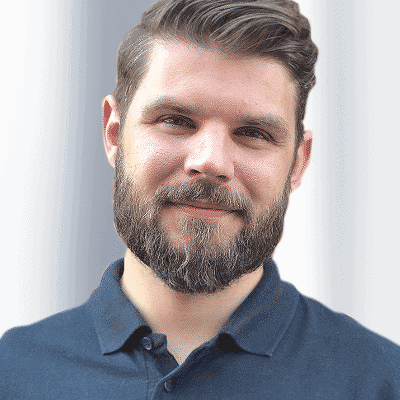 Philipp Greineder mit kurzen braunen Haaren und Bart, gekleidet in ein dunkles Polohemd, steht vor einem sanft verschwommenen Hintergrund mit Farbverläufen und präsentiert das souveräne Auftreten eines erfahrenen Referenten.