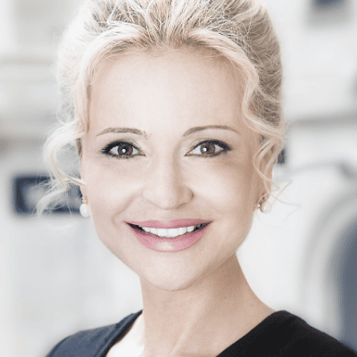 Sandra Navidi mit blondem, hochgestecktem Haar, leichtem Make-up und schwarzem Oberteil lächelt in einem sanft beleuchteten Innenraum in die Kamera und strahlt die ruhige Zuversicht eines erfahrenen Referenten aus.