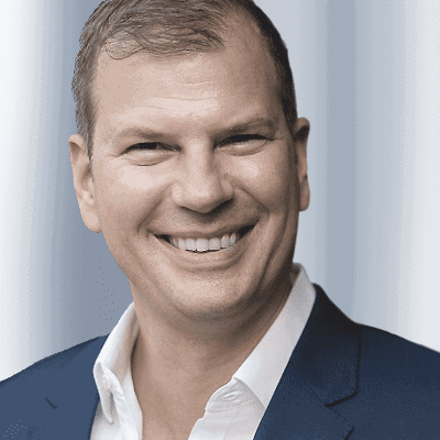 Wieland Arlt mit kurzen hellbraunen Haaren, der ein weißes Hemd und einen dunkelblauen Blazer trägt, lächelt in die Kamera vor einem unscharfen hellblauen Hintergrund - perfekt für die Verwendung als Referenten-Profilfoto.