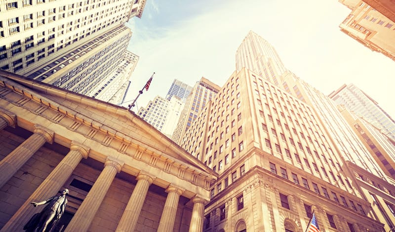 The tall skyscrapers and classic pillars of the financial district's buildings tower into the sky, symbolizing access to investment opportunities like fractional shares under a radiant sky.