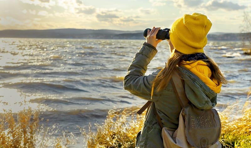 Eine Person mit gelber Mütze und grüner Jacke blickt durch ein Fernglas auf einen See mit Wellen und sucht die Landschaft ab wie eine Wertpapiersuche, mit Hügeln und einem bewölkten Himmel im Hintergrund.