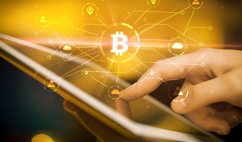 A hand interacts with a tablet displaying a glowing Bitcoin symbol and network connections representing digital cryptocurrencies, crypto trading and blockchain technology.