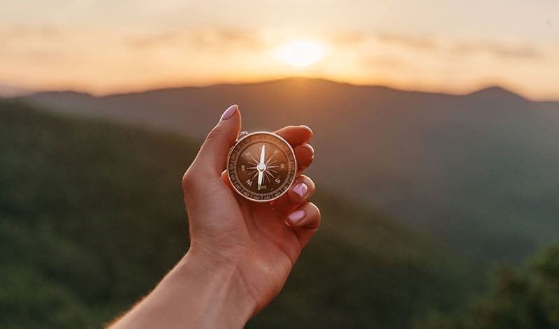 Im Vordergrund ist eine Hand zu sehen, die einen Kompass hält. Im Hintergrund sind Berge und die untergehende Sonne zu sehen, die SmartRouting symbolisieren, das Sie zu Ihrem Ziel führt.