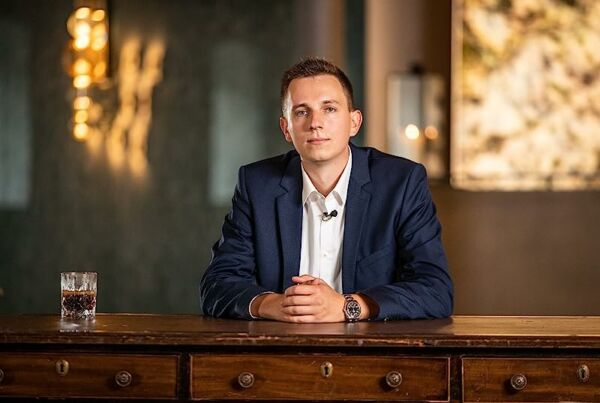 Moritz Hessel in einem marineblauen Anzug sitzt an einem Holztisch mit einem Glas eines dunklen Getränks und blickt in einem schwach beleuchteten Raum in die Kamera - ein perfektes Bild für jemanden, der über die totale Rückkehr nachdenkt.