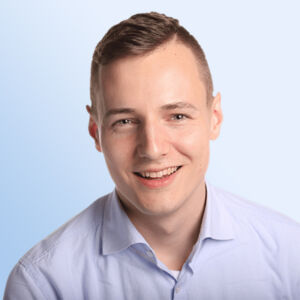 Aleks Bleck mit kurzen hellbraunen Haaren, der ein hellblaues Button-up-Hemd trägt, lächelt in die Kamera vor einem schlichten hellblauen Hintergrund.
