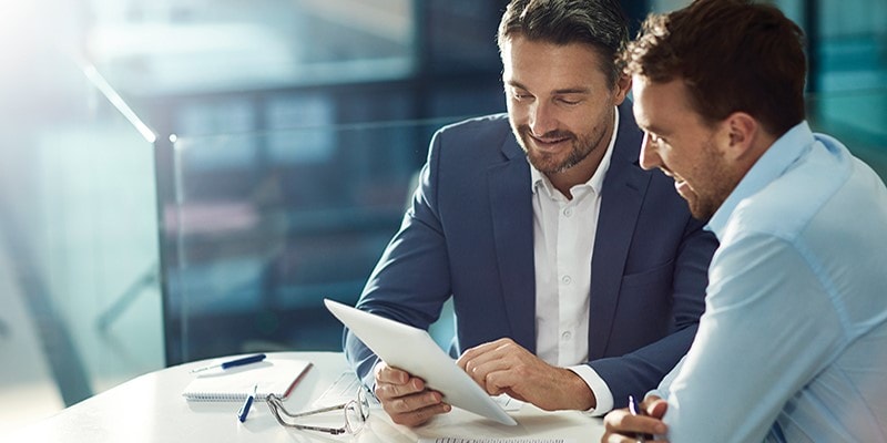 Zwei Männer in Geschäftskleidung sitzen an einem Tisch und betrachten gemeinsam ein digitales Tablet in einer hellen Büroumgebung, während sie über Managed Account-Optionen diskutieren.