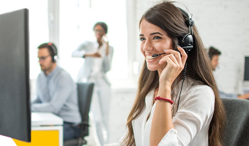 Eine Frau mit Headset sitzt an einem Schreibtisch und lächelt auf ihren Computer in einem Call Center, in dem das Quant4you Trading Terminal eingesetzt wird; andere Mitarbeiter arbeiten im Hintergrund.