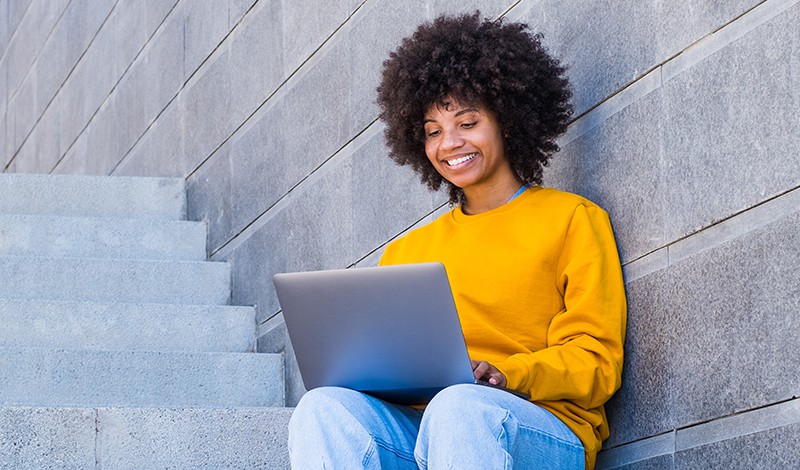 A person in a yellow sweatshirt sits on an outdoor staircase and smiles while using a laptop in front of a gray stone wall.