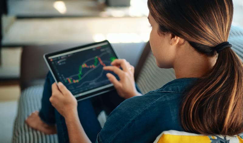 Eine Person sitzt auf einer Couch und hält ein Tablet in der Hand, auf dem ein Finanzdiagramm mit Candlestick-Grafiken und Marktdaten angezeigt wird.