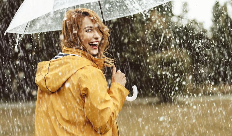 A person in a yellow raincoat holds a clear umbrella and smiles while standing in the pouring rain, unfazed by the storm - just as resilient as the people doing short sales - with blurry trees in the background.