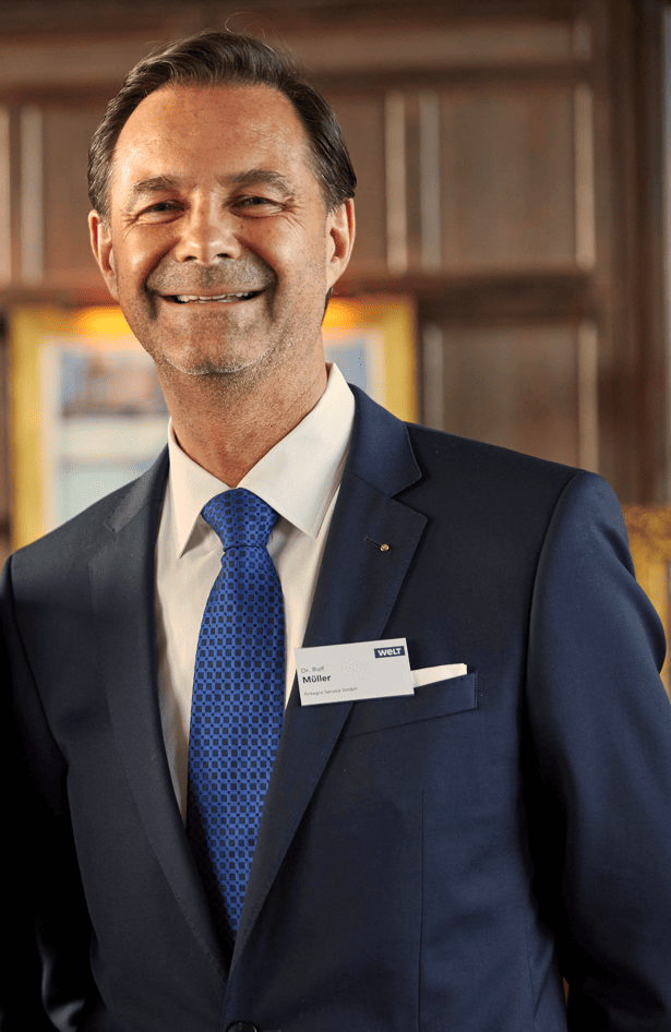 Dr. Rolf Müller in a blue suit and tie stands in a house and smiles at the camera. He is wearing a name tag on his jacket.