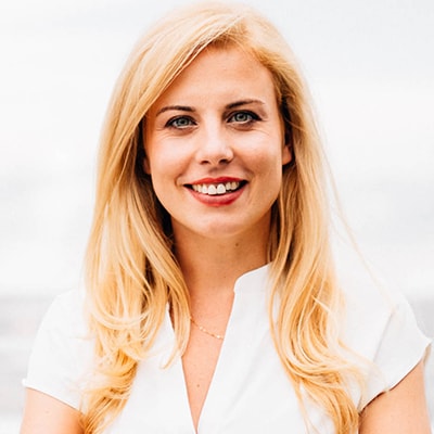 Dr Carmen-Mayer mit langen blonden Haaren, die ein weißes Oberteil trägt, lächelt in die Kamera vor einem hellen, unscharfen Hintergrund.