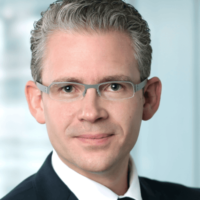Mario Steinrücken mit kurzen, gepflegten grauen Haaren, der eine Brille, einen dunklen Anzug, ein weißes Hemd und eine Krawatte trägt, posiert selbstbewusst vor einem unscharfen hellen Hintergrund - perfekt für professionelle Referentenprofile.