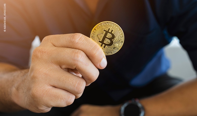 A person holds a gold-colored physical coin with the Bitcoin symbol, representing one of the many tradable products in the world of digital assets, against a blurred background.