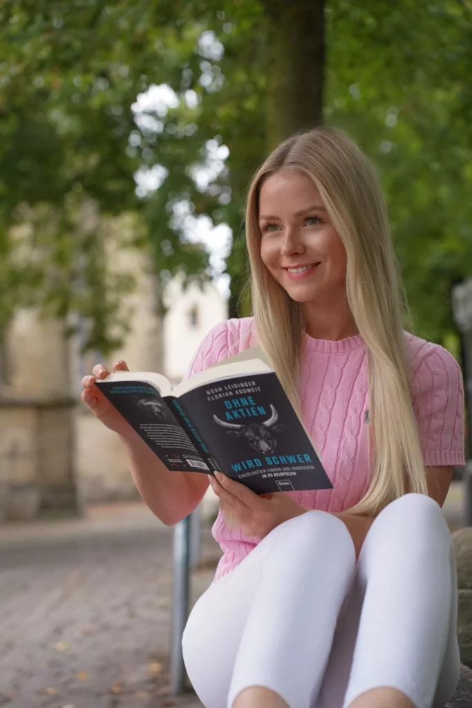 Eine Frau sitzt auf einer Bank und liest im Freien ein Buch mit der Aufschrift „Ohne Aktien wird schwer“ auf dem Cover.