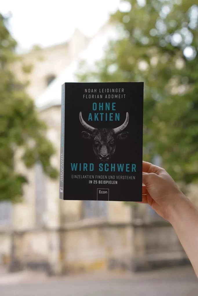 Eine Hand hält ein Buch mit dem Titel „OHNE AKTIEN wird SCHWER“ vor einem unscharfen Hintergrund hoch.