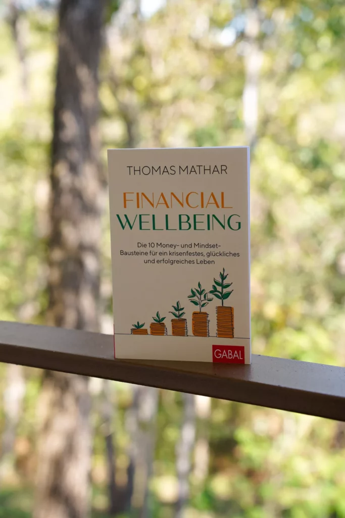 Ein Buch mit dem Titel „Financial Wellbeing“ von Thomas Mather, das sich auf Geld- und Denkstrategien für ein erfolgreiches und freudiges Leben konzentriert, ist vor einer natürlichen Kulisse mit Bäumen im Hintergrund platziert.