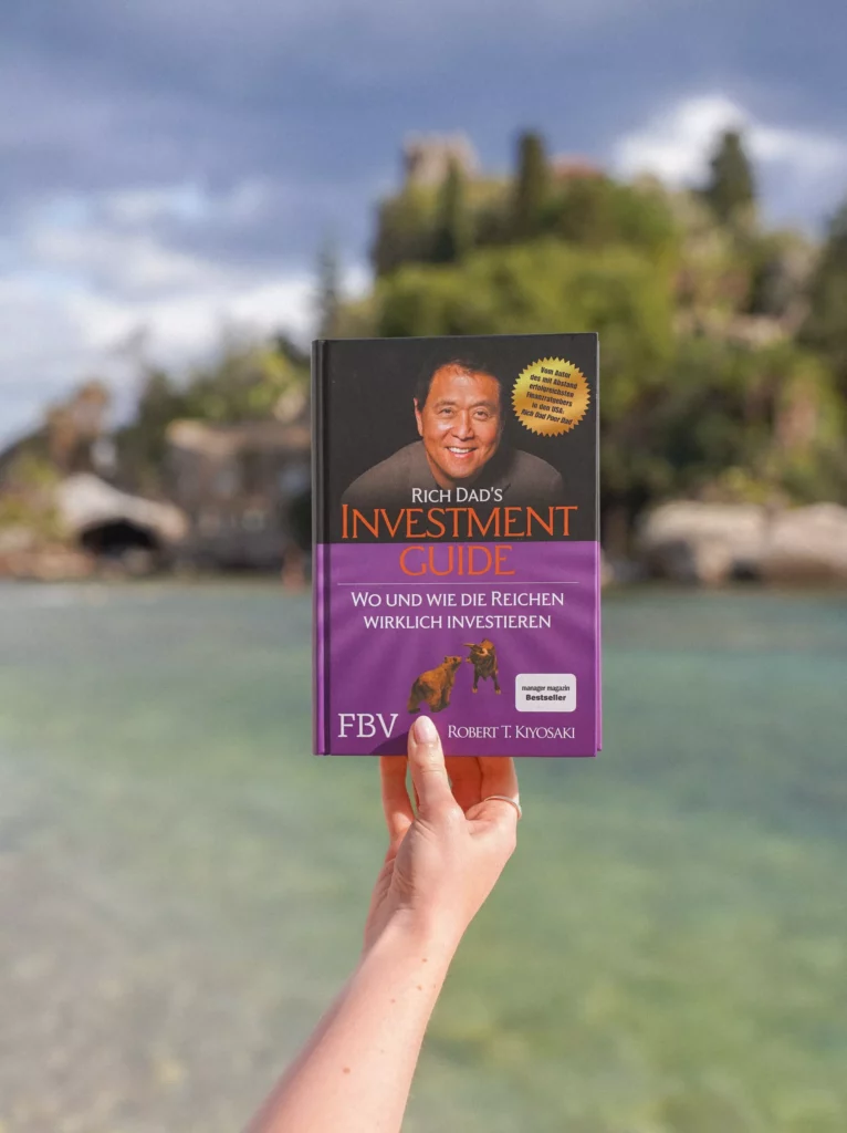 Eine Hand hält "Rich Dad's Investment Guide" von Robert T. Kiyosaki in die Höhe, wobei sich das Buch von einer sanft verschwommenen Szene im Freien mit Wasser und Grün abhebt.