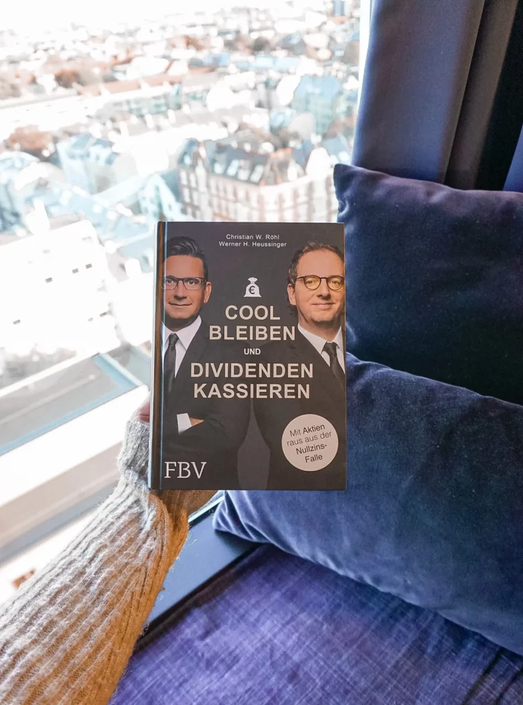 Eine Person hält ein Buch mit dem Titel „cool bleiben und Dividenden kassieren“ in der Hand, im Hintergrund eine urbane Skyline.