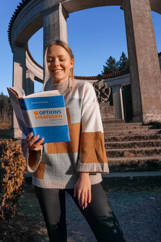 Frau lächelt und liest ein Buch mit dem Titel „Optionen Strategien“ in einer Außenumgebung mit klassischer Architektur im Hintergrund.